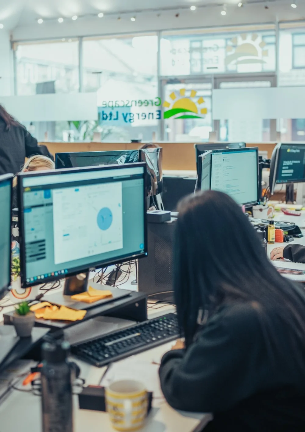 A Greenscape team member at her desk in the busy open-plan office, with the Greenscape Energy Ltd logo on the glass behind