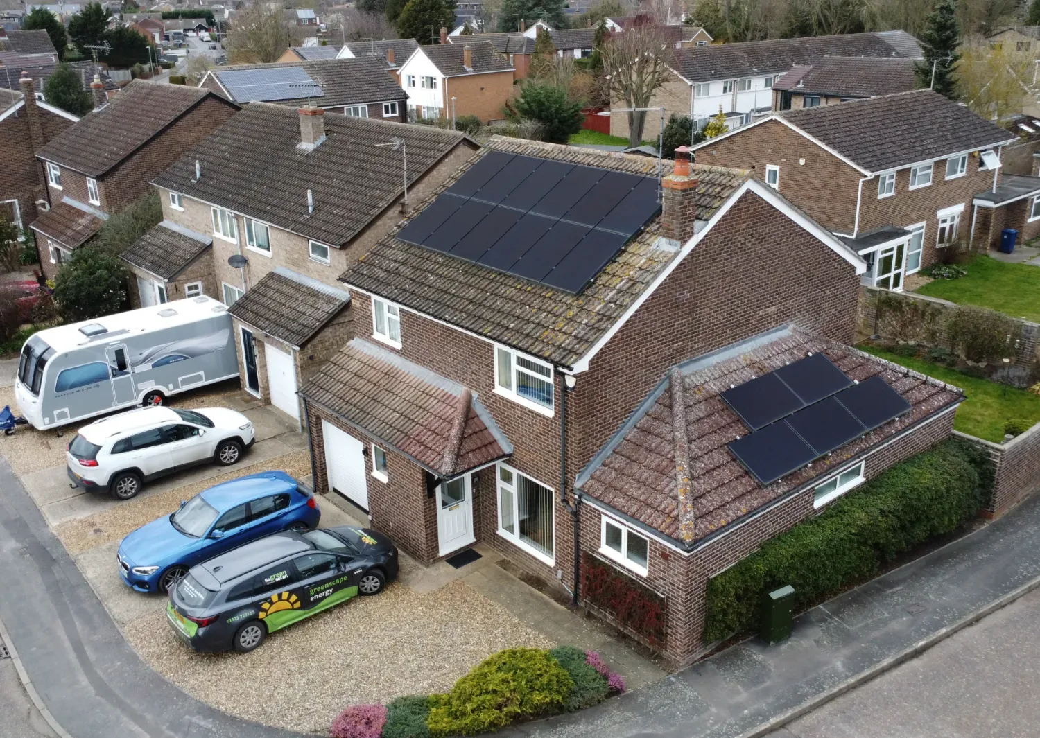 Aerial view of a brick semi-detached house with black solar panels on its main roof and garage, with a Greenscape car on the drive