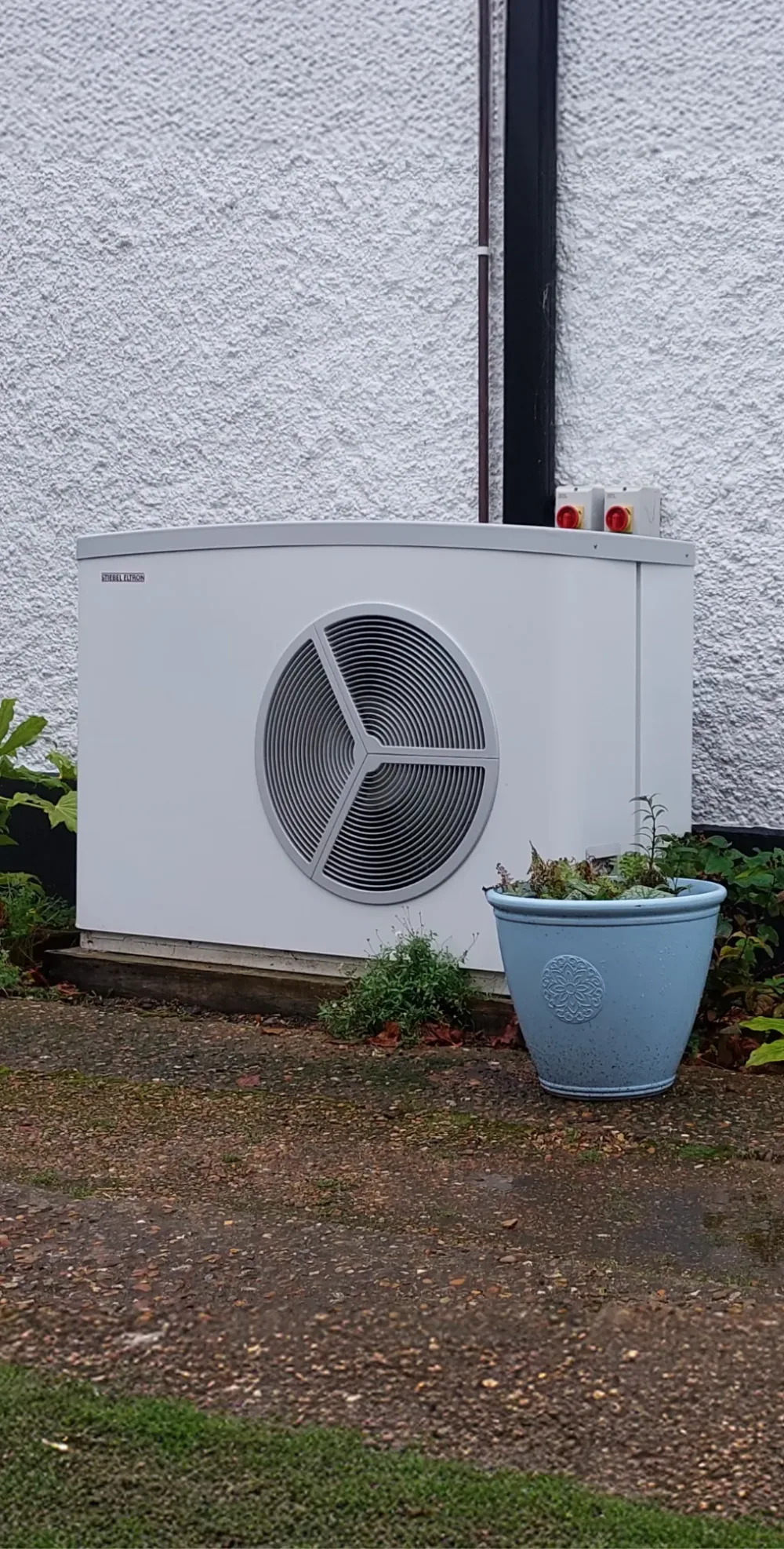 A Stiebel Eltron heat pump against a white rendered wall beside a blue plant pot