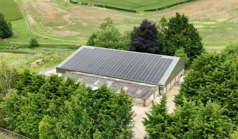 An aerial view of a green farm building with a full roof of solar panels, set among trees and fields