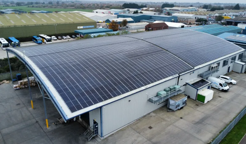 An aerial view of a curved industrial roof covered in solar panels, with coaches and units beyond