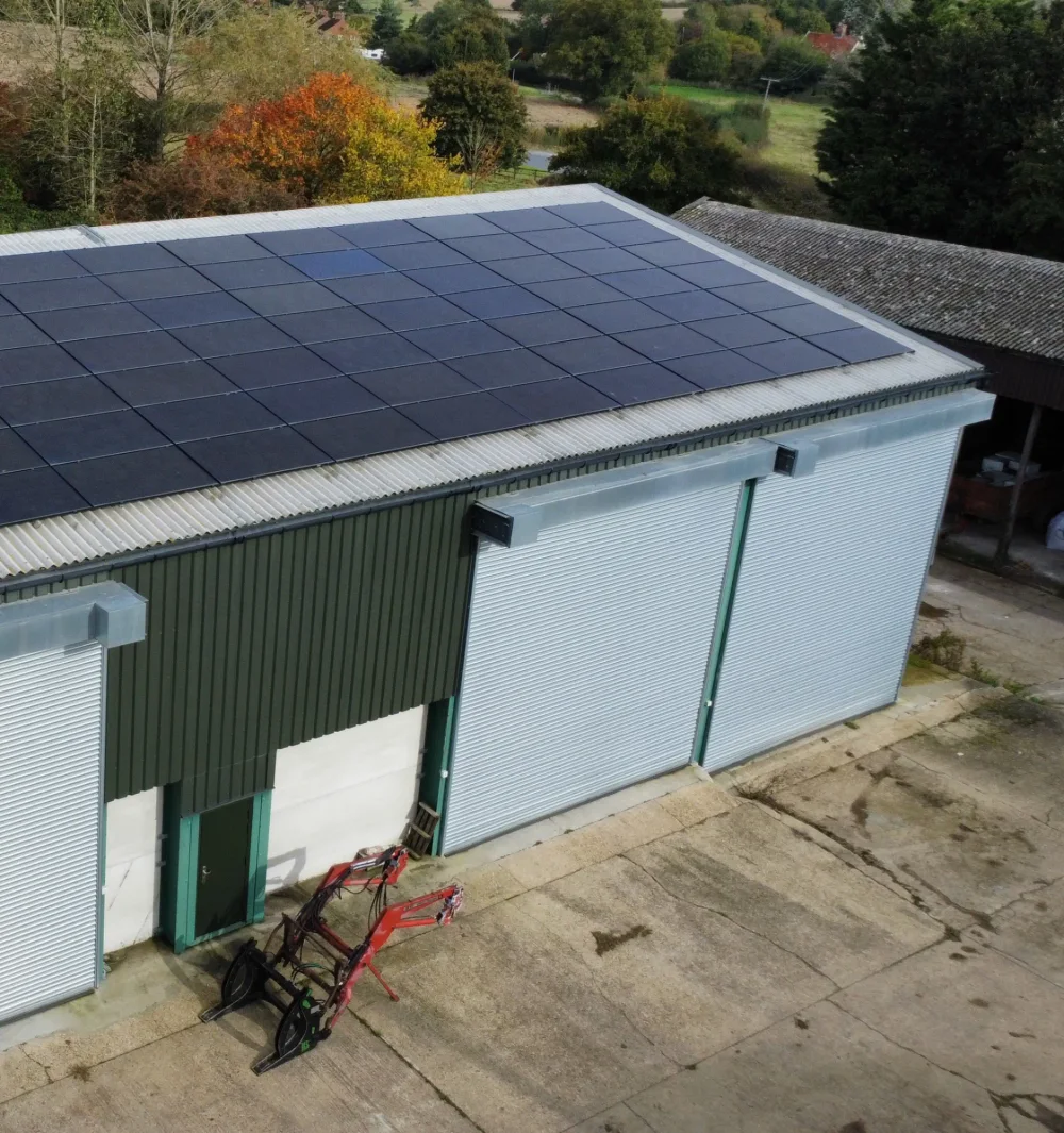 A large array of black solar panels on the roof of a green farm building, with a red loader attachment on the yard