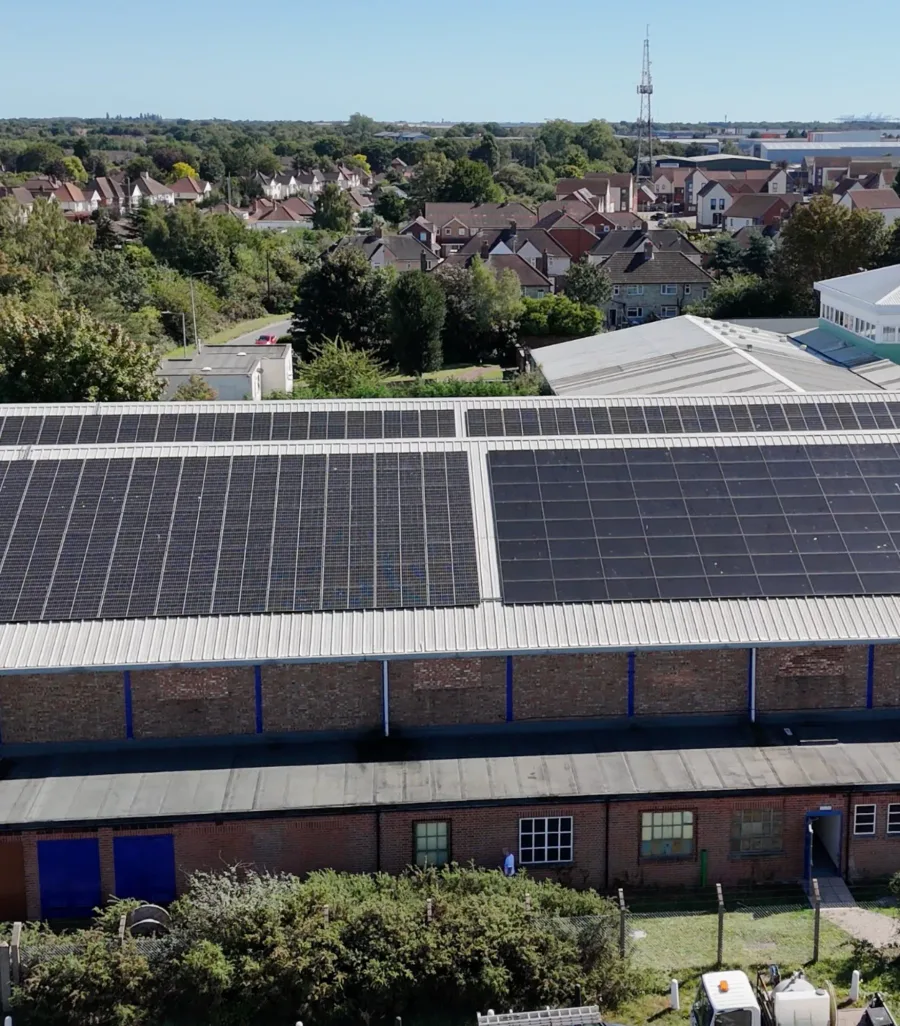 Large arrays of solar panels across a pair of industrial roofs, with houses and trees beyond