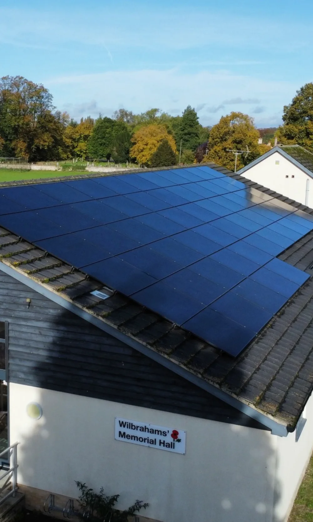 Black solar panels on the tiled roof of Wilbrahams' Memorial Hall, with autumn trees behind
