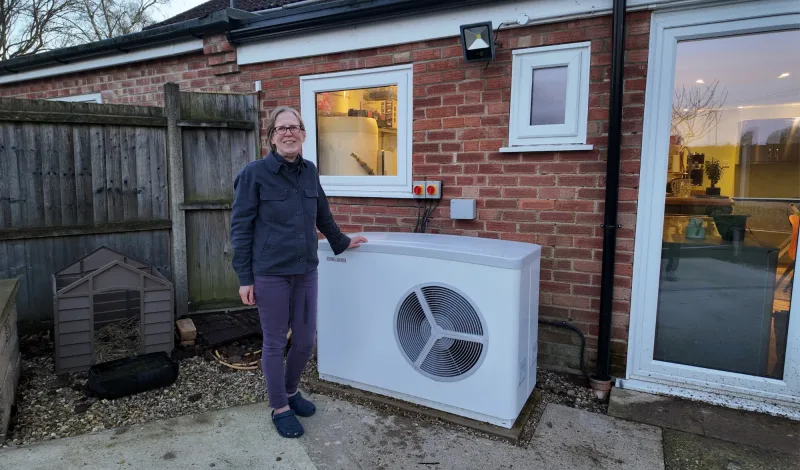 A woman smiling beside her white Stiebel Eltron heat pump against a red-brick wall