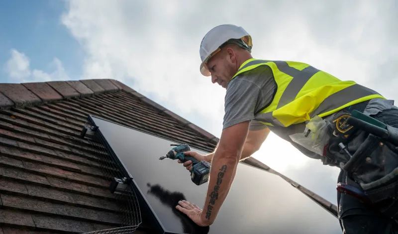 A Greenscape installer in a hard hat and hi-vis fixing a solar panel to a tiled roof