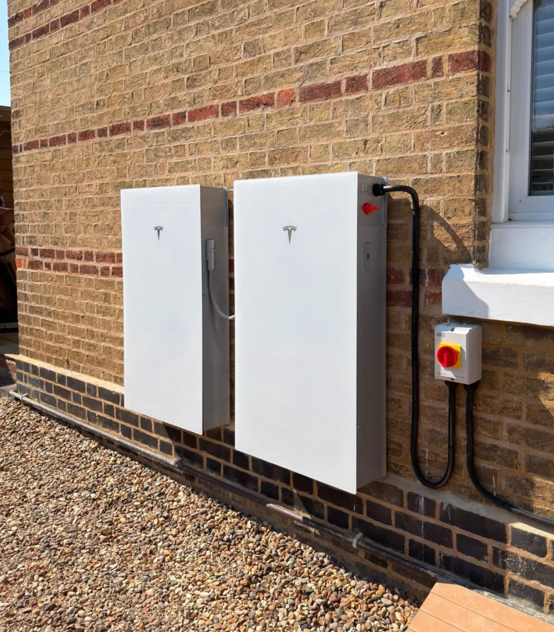 Two Tesla Powerwall batteries on a yellow-brick wall beside an isolator